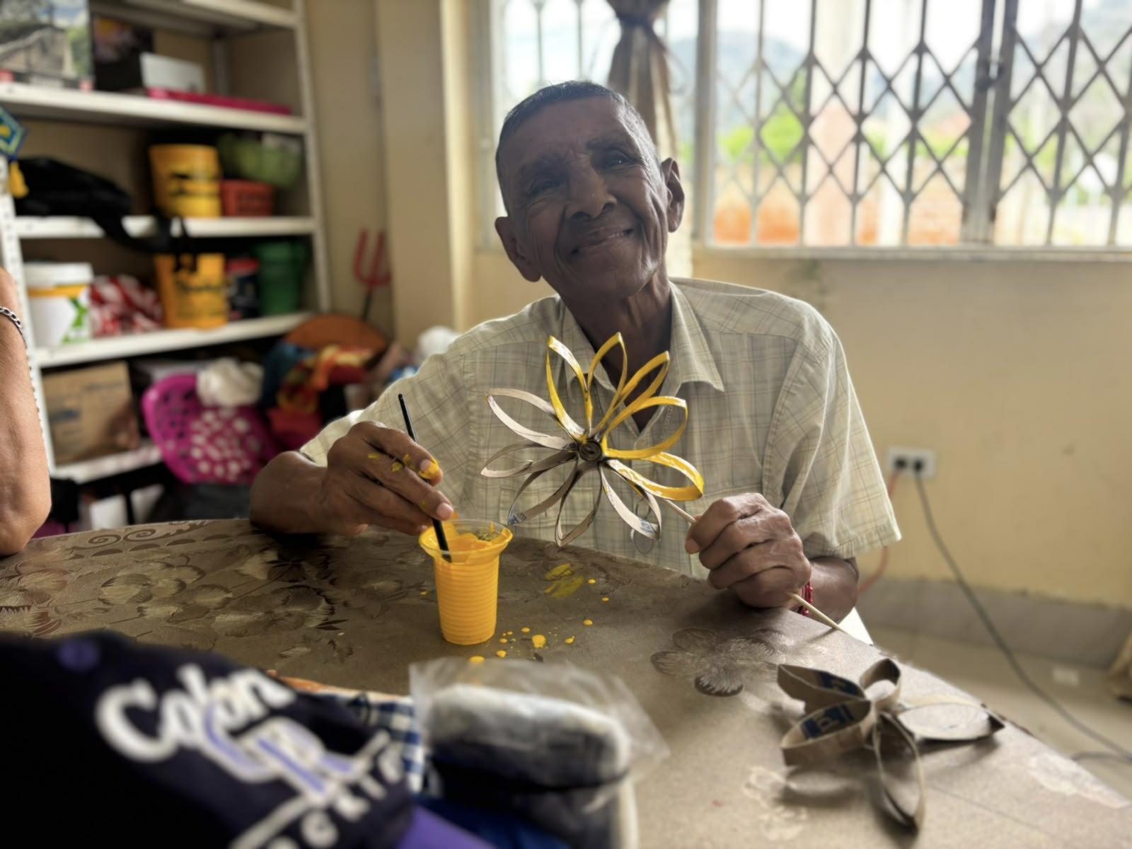 Un adulto mayor del Centro Diurno sonríe mientras pinta una flor hecha con material reciclado